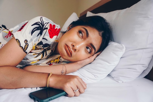 Triste joven frunciendo el ceño desde la cama esperando una llamada mientras sostenía el teléfono inteligente