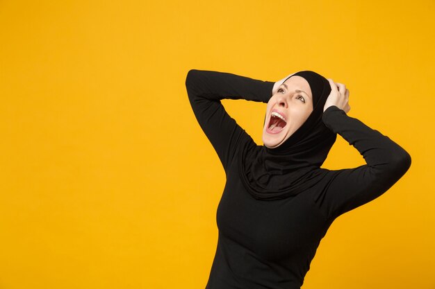 Triste, malestar, llanto, confundido, joven, árabe, musulmán, en, hijab, ropa negra, posar, aislado, en, amarillo, pared, portrait. Concepto de estilo de vida religioso del Islam de la gente.