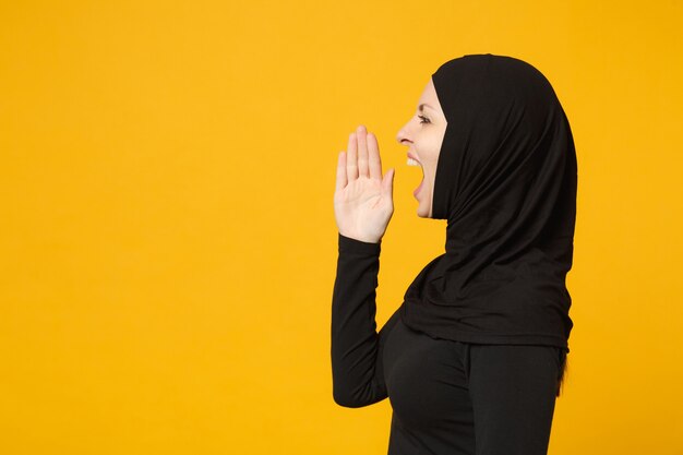 Triste, malestar, llanto, confundido, joven, árabe, musulmán, en, hijab, ropa negra, posar, aislado, en, amarillo, pared, portrait. Concepto de estilo de vida religioso del Islam de la gente.