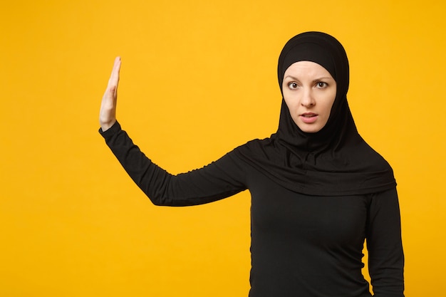 Triste, malestar, llanto, confundido, joven, árabe, musulmán, en, hijab, ropa negra, posar, aislado, en, amarillo, pared, portrait. Concepto de estilo de vida religioso del Islam de la gente.