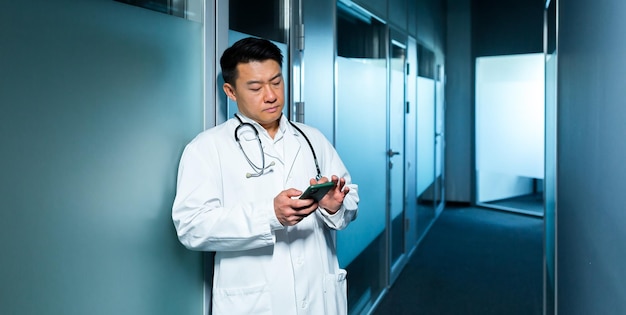 Triste médico asiático en el pasillo de la clínica usa el teléfono un hombre deprimido navegando por Internet en línea