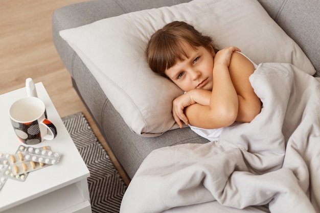 Triste menina doente e insalubre deitada sob um cobertor com tosse, olhando para a câmera, sentindo-se mal, tendo alta temperatura, posando sozinha no quarto, sente tristeza enquanto tem gripe.