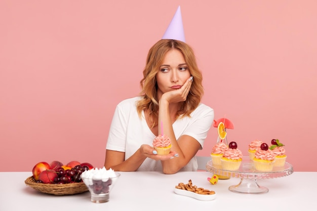 Triste mujer adulta joven con atuendo informal y cono de fiesta molesto porque sus invitados no vienen a su fiesta de cumpleaños sosteniendo un pastel con una vela en un estudio interior aislado en un fondo rosa