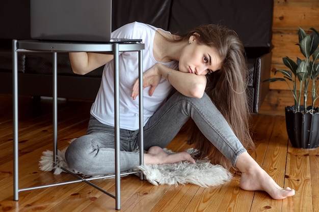 Triste mujer bonita con una computadora portátil mira pensativamente por la ventana y se aburre.