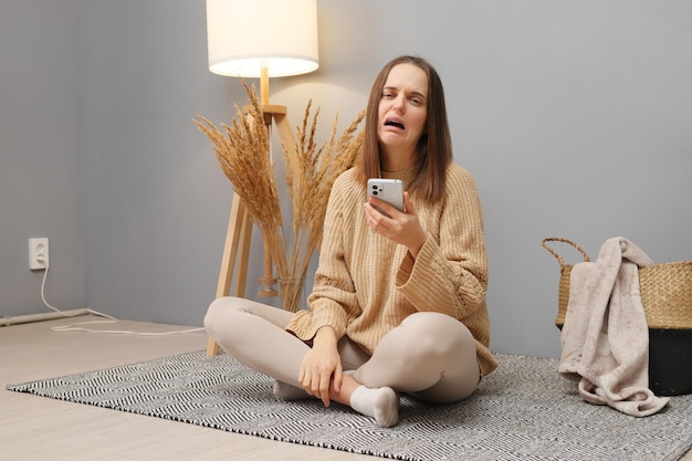 Triste mujer caucásica de cabello oscuro con ropa casual usando teléfono móvil y llorando no tiene conexión a Internet o la batería se acabó en el momento equivocado sentada en el suelo en el interior de la casa