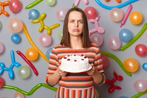 Triste mujer decepcionada con cabello castaño con vestido a rayas sosteniendo pastel que no le gusta mezclar orden comprando postre equivocado de pie contra la pared gris decorada con globos de colores