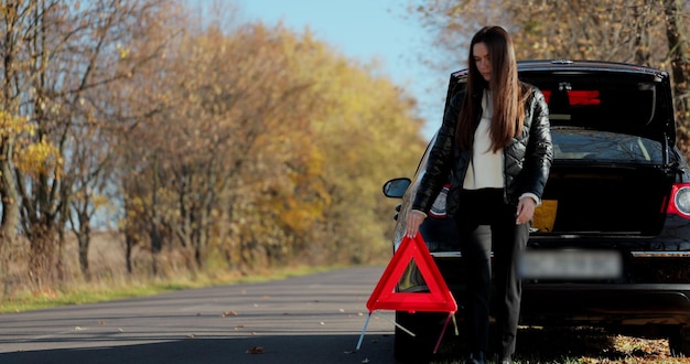 Triste mujer decepcionada en coche roto. Conductora de mujer instalando el letrero de triángulo rojo. Accidente de coche roto. Mujer conductora coloca señal de parada de emergencia.