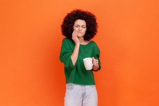 Triste mujer enferma con peinado afro con suéter verde de estilo casual tiene dolor de dientes después de beber bebidas frías o calientes, lesión dental. Disparo de estudio interior aislado sobre fondo naranja.