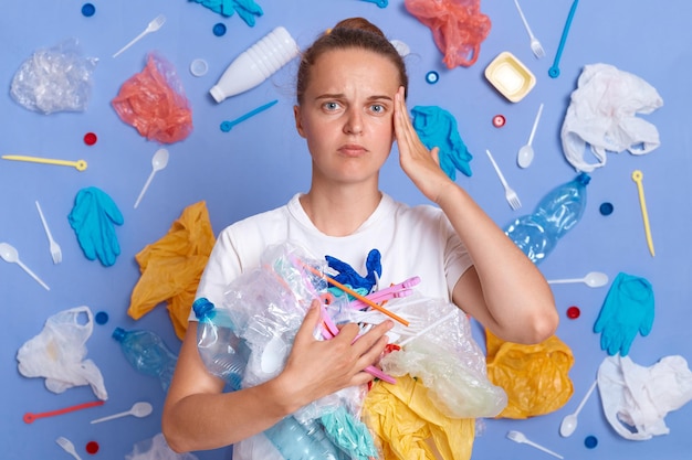 Triste mujer infeliz con camisa blanca aislada en la pared azul con basura alrededor del territorio de limpieza de basura mirando a la cámara con expresión disgustada