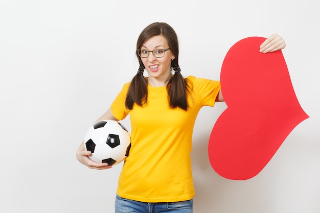 Triste mujer joven europea, colas de caballo, aficionado al fútbol o jugador con gafas, uniforme amarillo mantenga bola clásica, gran corazón rojo aislado sobre fondo blanco. Deporte, fútbol, concepto de estilo de vida saludable.