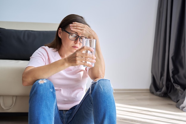 Triste mujer de mediana edad sentada en casa en el suelo con un vaso de agua