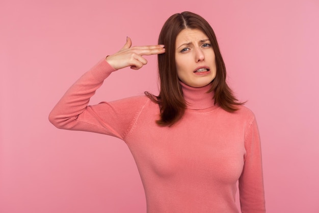 Triste mujer morena frustrada sosteniendo los dedos cerca de la cabeza imitando el arma mirando a la cámara con expresión estresada de pánico Disparo de estudio interior aislado sobre fondo rosa