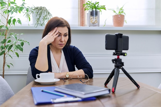 Triste mujer de negocios madura en casa usando un teléfono inteligente para videoconferencia