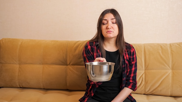 Foto triste mujer de pelo largo en camisa a cuadros recoge el agua que fluye desde el techo hacia la olla sentada en un cómodo sofá en la sala de estar moderna