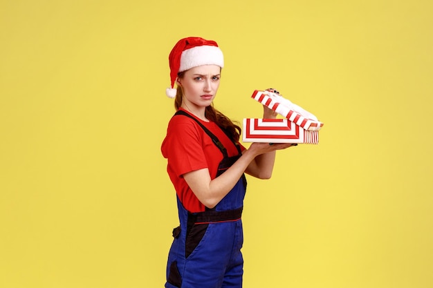 Triste mujer trabajadora molesta sosteniendo una caja envuelta abierta, decepcionada del regalo que recibe, usando un mono azul y un sombrero de santa claus. Disparo de estudio interior aislado sobre fondo amarillo.