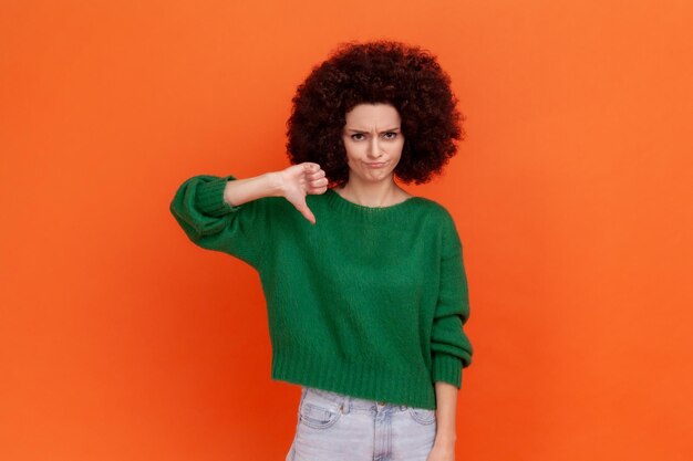 Triste mulher deprimida com penteado afro vestindo suéter verde estilo casual mostrando o polegar para baixo mau serviço ou conteúdo não recomenda tiro de estúdio interior isolado em fundo laranja