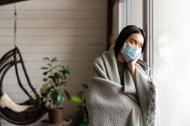 Triste niña asiática enferma en cuarentena de pie cerca de la ventana en casa y mirando afuera vistiendo ropa médica ...