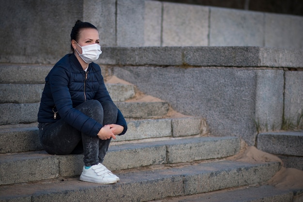 Triste niña deprimida molesta, joven y solitaria mujer frustrada sentada en las escaleras, sufriendo debido al aislamiento, coronavirus. Persona en máscara protectora médica en cara. Corazón roto, virus, concepto epidémico