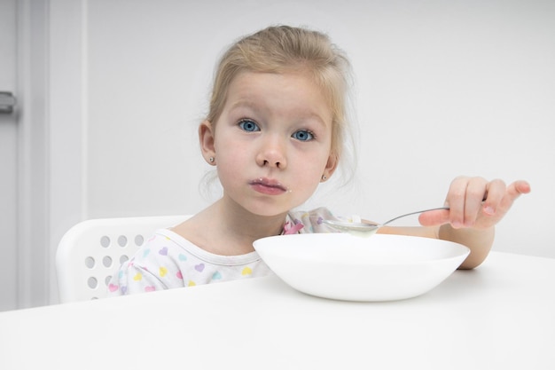 Triste niña rubia come gachas de avena en una mesa blanca