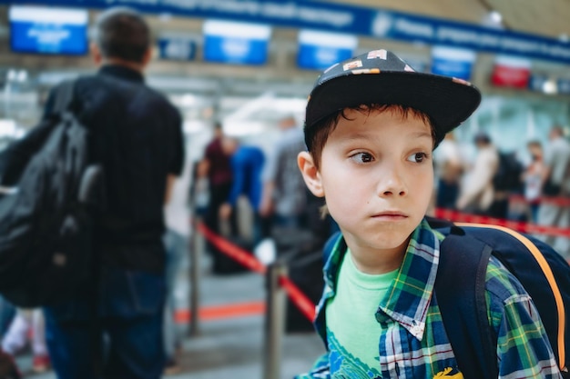 Triste niño caucásico solo en el aeropuerto con mochila y maleta en el puesto de facturación esperando en la fila