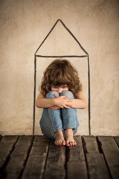 Triste niño sin hogar siiting en el suelo en una habitación oscura