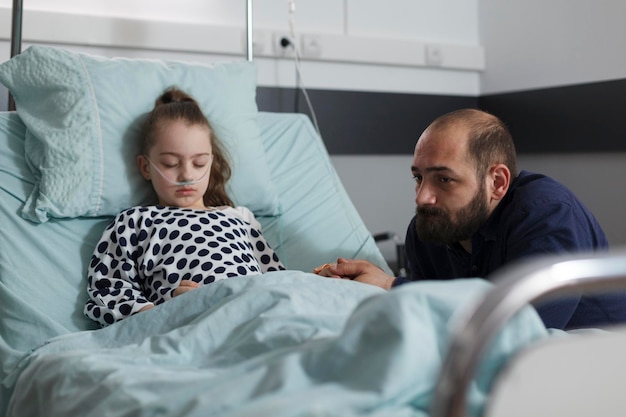 Foto triste padre sentado junto a una niña enferma hospitalizada descansando en la cama de un paciente dentro de la sala de pediatría del hospital. niño pequeño enfermo con tubo nasal descansando mientras los padres esperan despertarse de la anestesia.