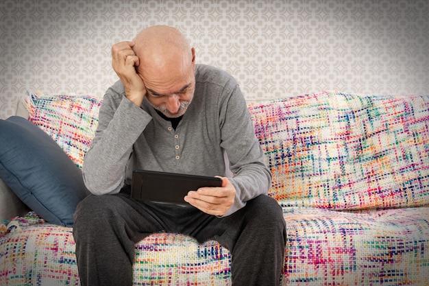 Triste y pensativo anciano sentado en el sofá con Tablet PC en la mano