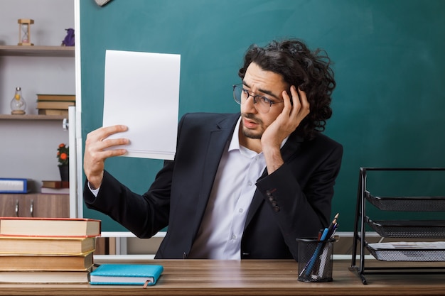 Triste profesor con gafas sosteniendo y mirando el papel sentado a la mesa con herramientas escolares en el aula