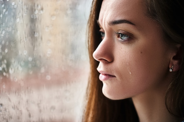 Foto triste triste mulher chorando com lágrimas olhos que sofrem de choque emocional, perda, tristeza, problemas de vida e romper o relacionamento perto da janela com pingos de chuva. mulher recebeu más notícias