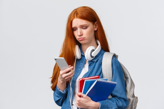 Triste y triste niña haciendo pucheros con el pelo rojo y astuto, frunciendo el ceño suspirando y mirando con arrepentimiento en la pantalla del teléfono inteligente