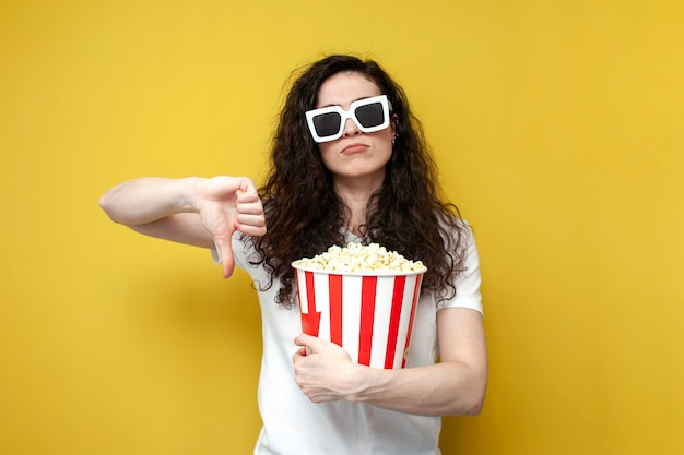 Triste visor de niña infeliz en gafas 3d sosteniendo palomitas de maíz y mostrando aversión sobre fondo amarillo