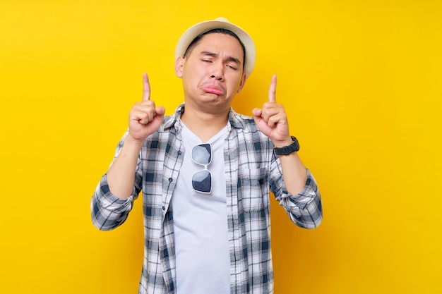 Tristeza Joven apuesto hombre de etnia asiática de 20 años con ropa informal sombrero señalando con el dedo por encima de la cabeza indicando en el espacio de trabajo maqueta del espacio de copia aislada en fondo amarillo Concepto de estilo de vida de la gente