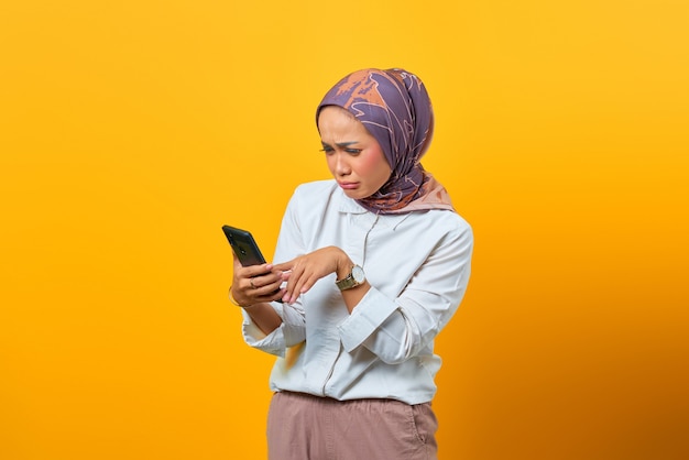 Tristeza mujer asiática mirando smartphone recibiendo malas noticias sobre fondo amarillo