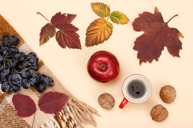 Trockene gelbe und braune Blätter, Tasse Kaffee, Walnüsse und kariertes Plaid mit Trauben auf beigem Hintergrund. Eine Herbstkomposition. Ansicht von oben.