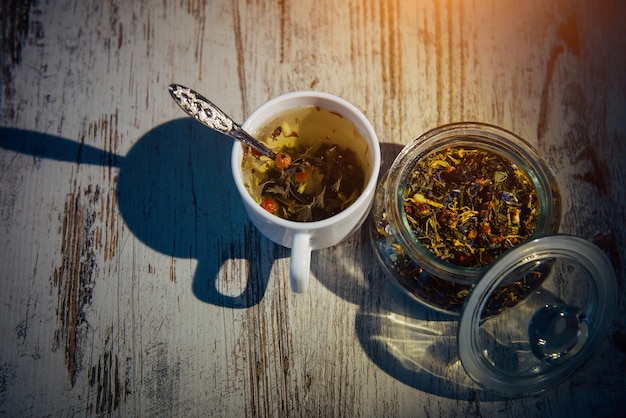 Trockene Mischung aus Teeblättern, Kräutern, Blütenblättern, Beeren und Nüssen in Glasschüssel. Natürlicher Kräutertee in weißer Tasse auf grauem Holzhintergrund im Abendlicht, Schatten auf dem Tisch.