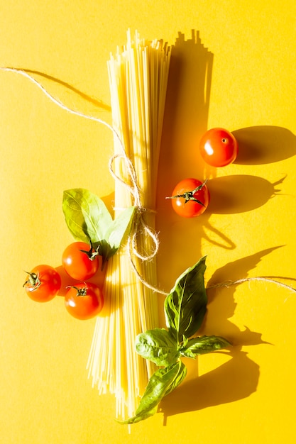 Trockene Spaghetti auf einem gelben Hintergrund neben Kirschtomaten und einem Basilikumblatt