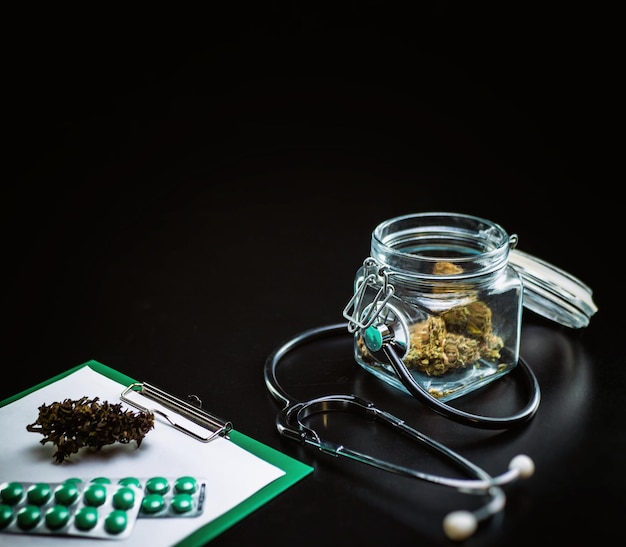 Trockenes medizinisches Cannabis in einem Glas mit einem Stethoskop auf schwarzem Hintergrund