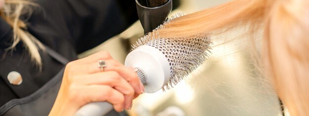 Trocknen von glattem blondem Haar mit schwarzem Haartrockner und weißer Rundbürste im Friseursalon aus nächster Nähe