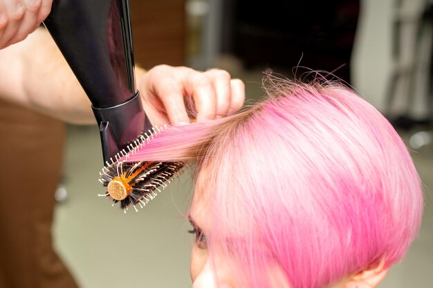 Trocknen von kurzen rosa Haaren einer jungen kaukasischen Frau mit einem schwarzen Haartrockner und einer schwarzen Rundbürste durch die Hände eines männlichen Friseurs in einem Friseursalon aus nächster Nähe