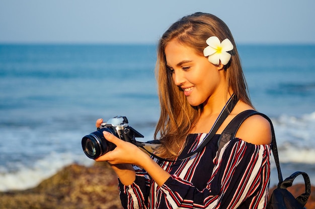 Tropischer Genuss Sommertourismus und Fotografie. Junge reisende Frau in einem stylischen gestreiften Overall mit Kamera und Rucksack in der Nähe von Meerstrand Indischer Ozean Goa türkisfarbenes Wasser, Himmel und Felsen.