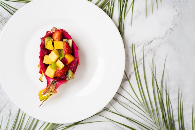 Tropischer Obstsalat serviert in halber Drachenfrucht. Ansicht von oben. Sommersalat auf weißem Teller auf dem Tisch. Gesundes Frühstück, Lebensmittelkonzept. Flach liegen. Vertikale Aufnahme.