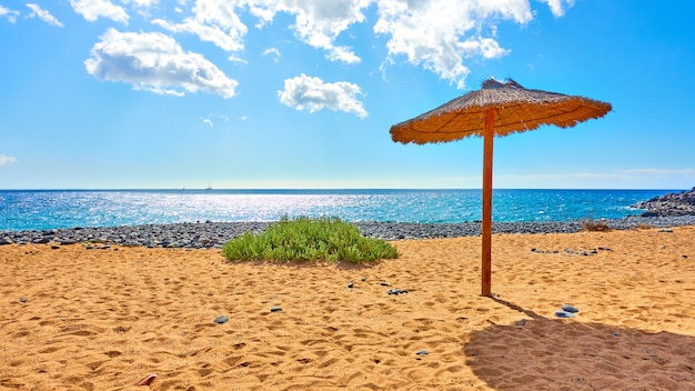 Tropischer Strand mit Strohsonnenschirm am Meer an einem sonnigen Sommertag
