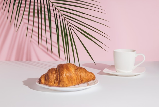 Tropisches Palmblatt-Croissant und eine Tasse Kaffee auf rosa Hintergrund Minimales Essenskonzept Frühstück