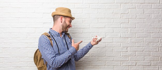 Trouxa vestindo do homem novo do viajante e uma câmera do vintage que aponta ao lado, sorrindo surpreendido apresentando algo, natural e ocasional. Segurando um modelo de avião.