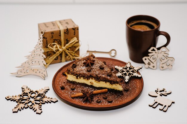 Un trozo de delicioso pastel de chocolate con crema, granos de café en un plato marrón, taza de café se aísla en una superficie blanca. Canela, anís. Regalos y adornos navideños. Vista lateral.