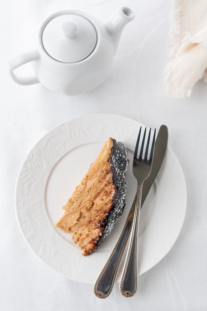 Un trozo de pastel de galletas multicapa hecho en casa Empapado en crema de mantequilla Adornado con glaseado de chocolate y coco En un plato blanco con un tenedor y un cuchillo Primer plano Fondo blanco Vista desde arriba