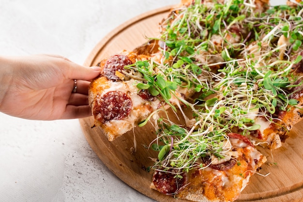 Trozo de pinsa romana en mano con estiramiento de mozzarella con salami, queso, champiñones, decorado con microgreens sobre tabla de madera.