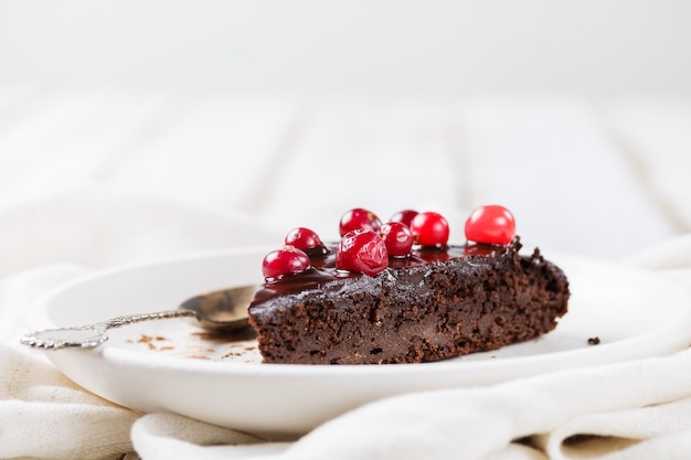 Un trozo de tarta de brownie sin gluten con glaseado de chocolate, decorado con arándanos, sobre una placa blanca, sobre una mesa de madera clara de cerca. Fondo claro borroso. Postres Saludables.