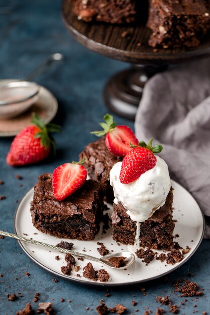 Trozos de brownie de nueces de chocolate, con bola de helado, sobre plato blanco, cuchara, con rodajas de fresas, migas, textil gris. Fondo azul oscuro. Vertical