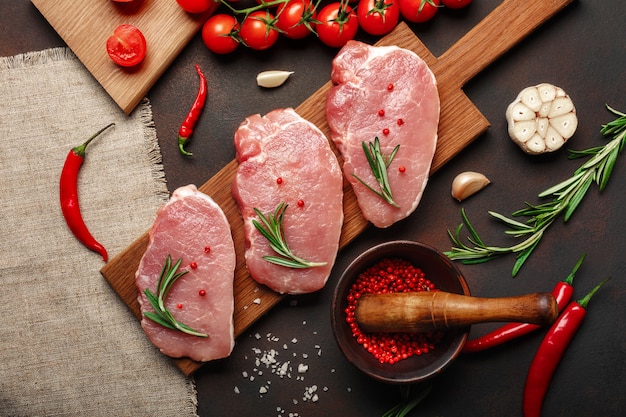 Trozos de carne cruda de cerdo en tabla para cortar con tomates cherry, romero, ajo, pimienta, sal y especias, mortero